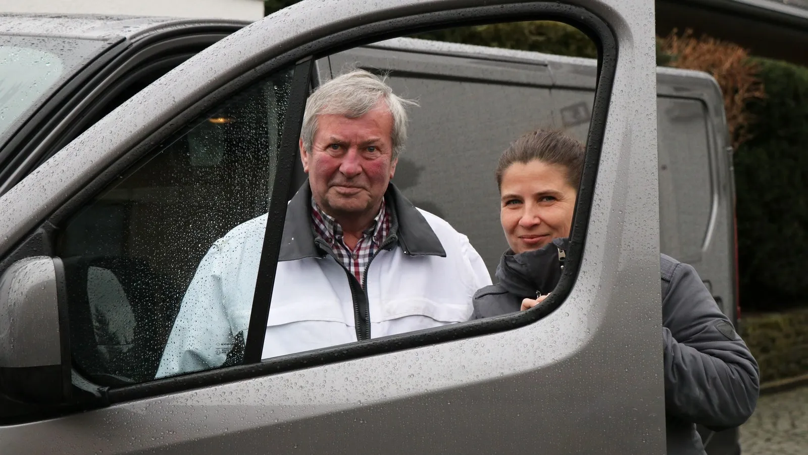Ein Mann und eine Frau stehen hinter einer geöffneten Autotür und schauen durch das Fenster in die Kamera.